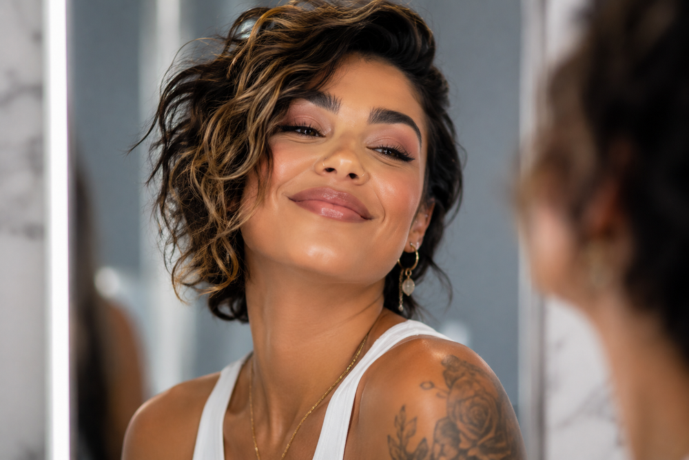 A close-up lifestyle shot of a woman with short, wavy brown hair looking into a mirror, wearing a white tank top and gold jewelry.