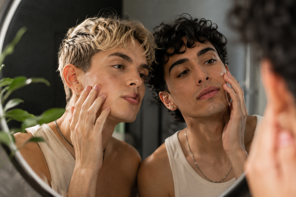 Two young men looking at their reflections in a round bathroom mirror, touching their faces. One has short blonde hair and the other has curly dark hair, both wearing light beige tank tops.